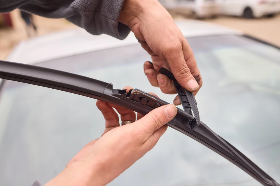 Personne installant un essuie-glace neuf sur le pare-brise d'un v&eacute;hicule, manipulation des clips de fixation avec les deux mains, op&eacute;ration de maintenance automobile en ext&eacute;rieur
