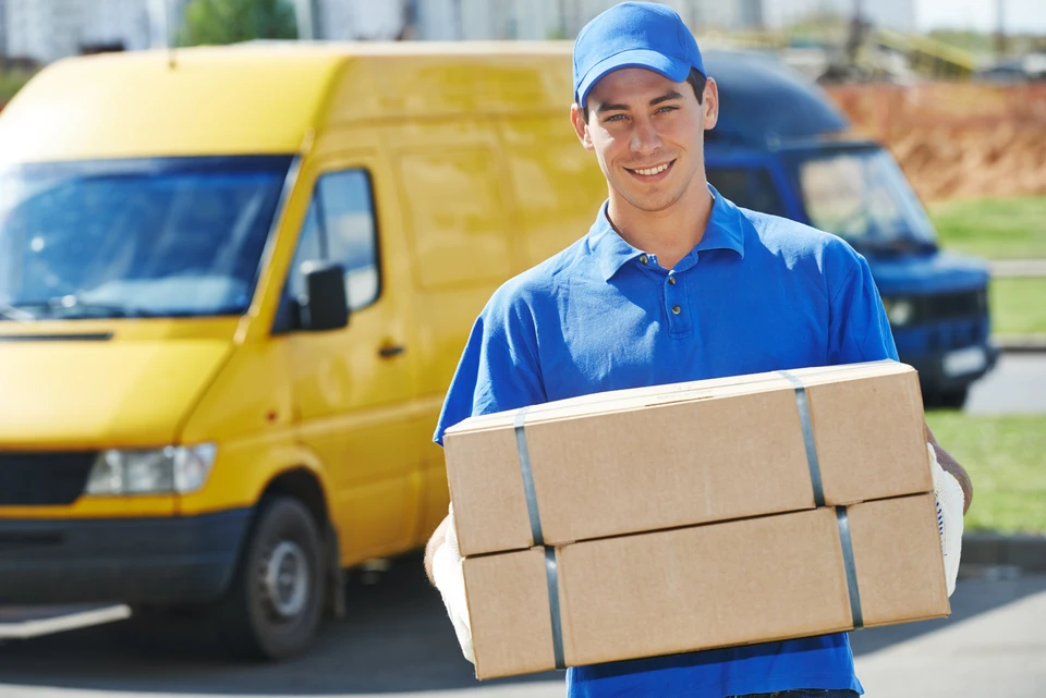 Livreur souriant en uniforme bleu et casquette portant des colis emballés devant un camion de livraison jaune stationné dans un environnement résidentiel ensoleillé