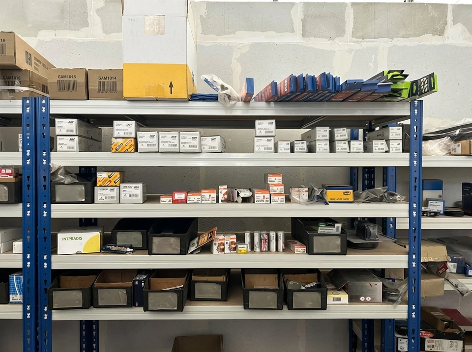 Un distributeur de pièces détachées automobiles en tenue professionnelle dans son magasin, consulte une base de données ou un catalogue technique sur un écran en arrière-plan, tenant une pièce détachée automobile (moteur, alternateur, ou autre composant standard) dans ses mains. L'environnement doit montrer des étagères organisées de pièces automobiles en arrière-plan, symbolisant l'expertise technique et la connaissance approfondie du secteur. La scène doit transmettre la professionnalité, la compétence et l'approche conseil personnalisée.